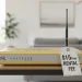 An outdated beige ISP router sits in a modern living room with a prominent '$15/mo RENTAL FEE' price tag tied to its antenna.